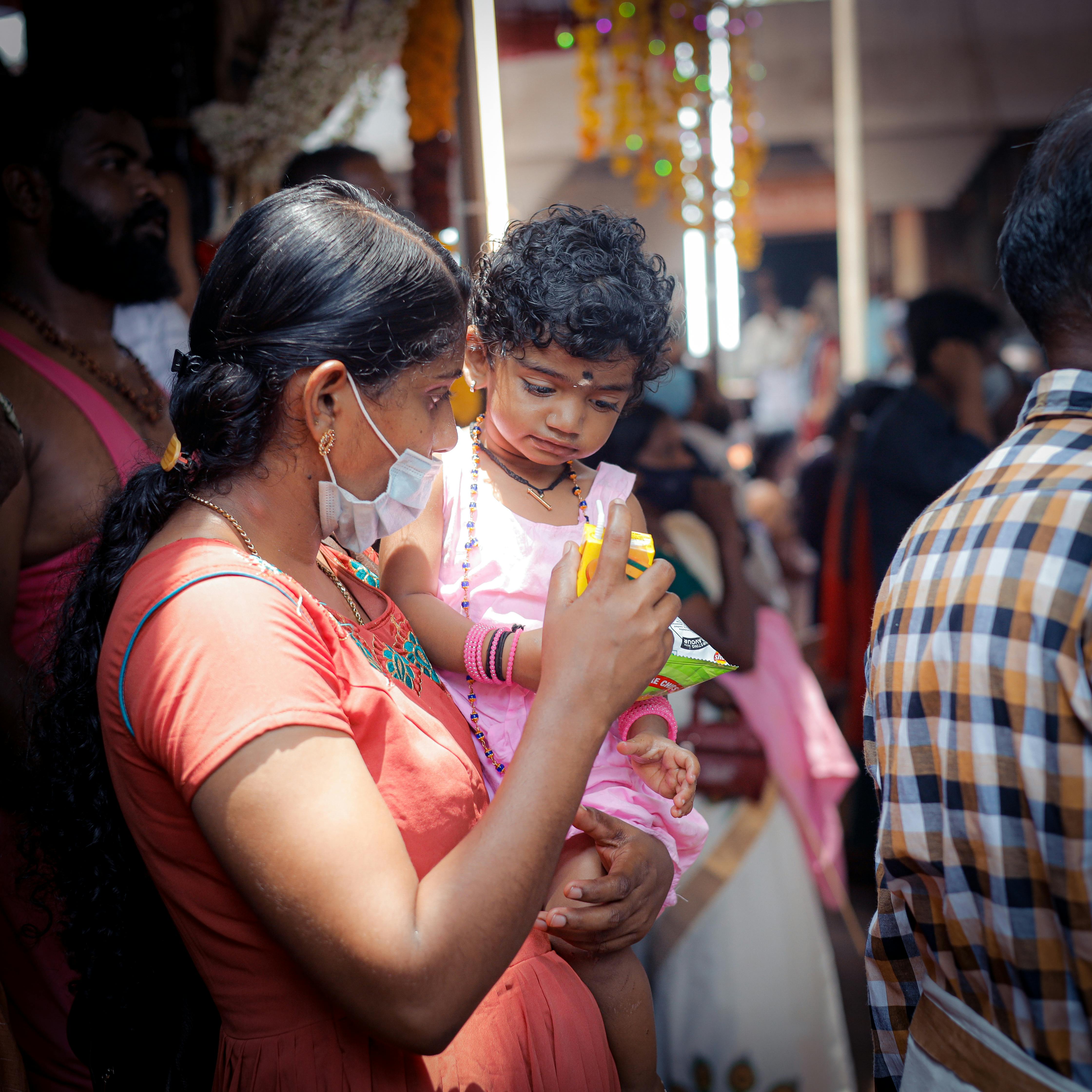A mother holds her child during an outdoor cultural event, surrounded by people.